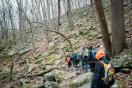 A group of hikers, wearing colorful jackets and backpacks, walks along a rocky forest trail during a cool, overcast day. The forest is dense with bare trees and moss-covered rocks scattered across the ground. The atmosphere suggests early spring or late autumn. The hikers appear to be navigating a slightly steep descent.